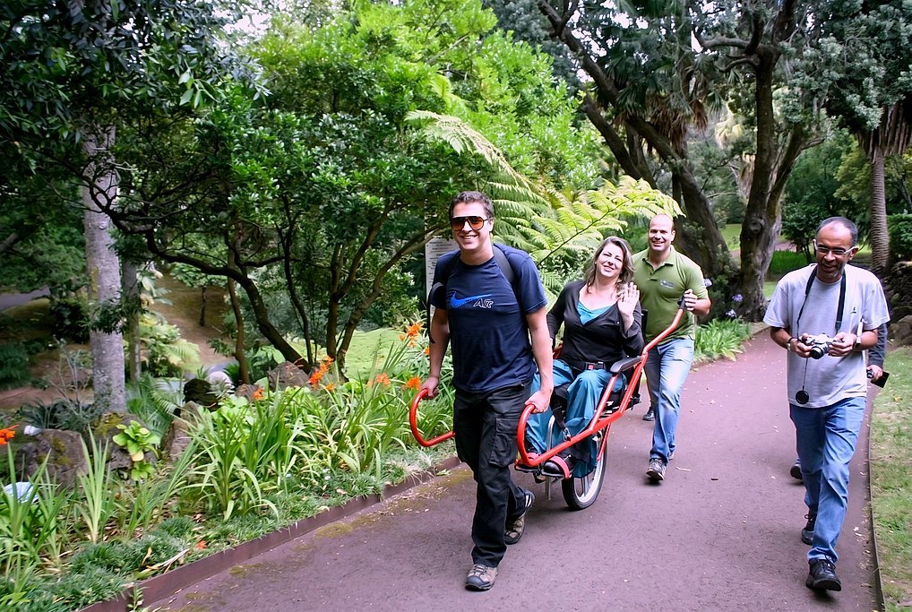 Two monitors guide a smiling seated rider in the Joelette chair along a garden path with orange tropical flowers.