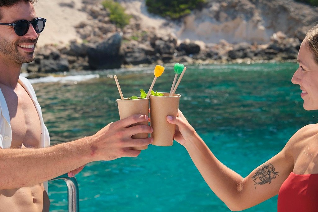 Man and woman toasting with drinks in paper cups near a rocky coastline.